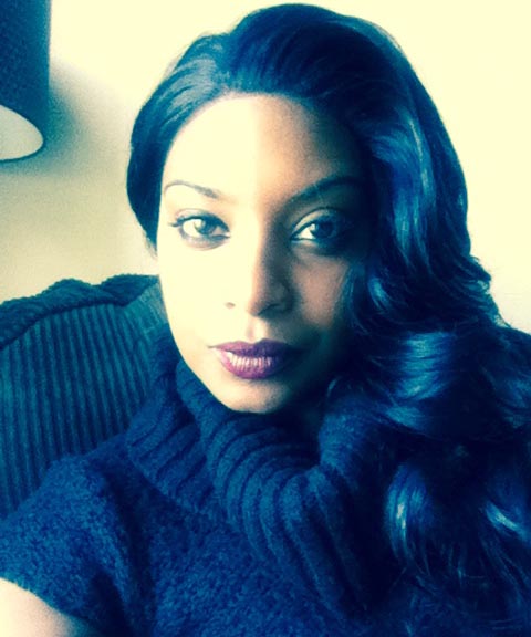 Image description: Kenya Lowe, a black woman with long black hair, wearing a black knitted sweater top with short sleeves. There appears to be a lamp with brown shade in the corner of the photo. She is sitting in a brown chair.  She has a slight smile.